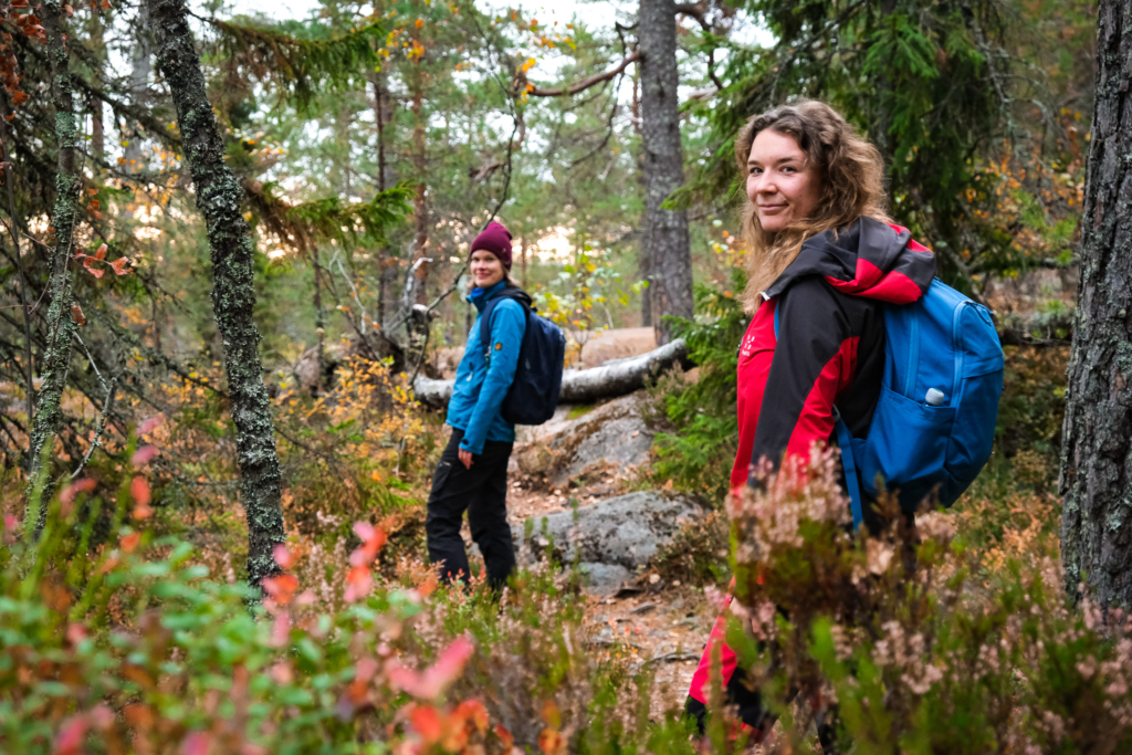 Nuuksioruska Pääkaupunkiseutu tarjoaa hienoja metsiä: Siniseen ja punaiseen takkiin pukeutuneet naiset kävelevät syksyisessä metsässä ja katsovat kohti kameraa. Sinäkin voit saada hyvinvointia omaan elämään metsästä.