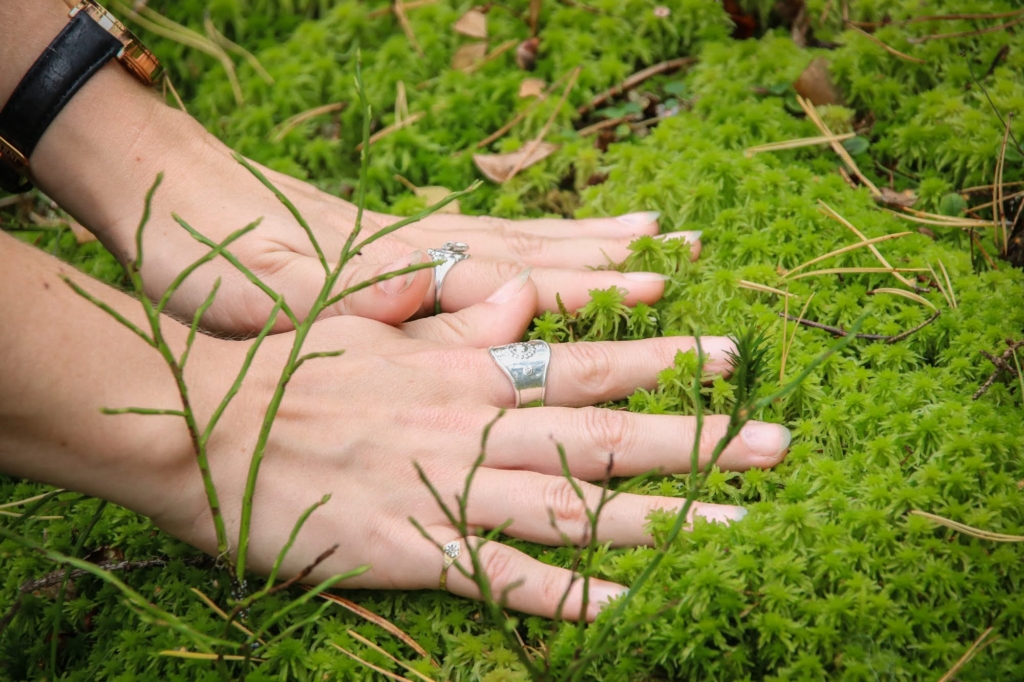 Hyvinvointia työhön. Kuvassa kädet, joissa kummassakin on yksi hopeinen sormus, painavat hellästi pehmeän näköistä, vaaleanvihreää rahkasammalmätästä. Sammalella on keltaisia männynneulasia ja lehdettömiä mustikanvarpuja.