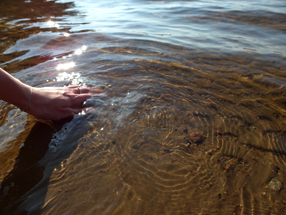 Hyvinvointia työhön. Hiekkapohja kuultaa kirkkaan veden lävitse, ja kuvan vasemmassa reunassa käsi koskettaa vettä, saaden sen pinnan väreilemään ja välkehtimään auringossa.